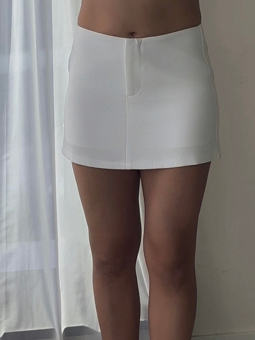 Person wearing a white skirt and white high-heeled shoes in a room with a curtain.